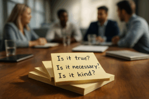 A stack of yellow sticky notes sits on a conference table in sharp focus, each note displaying the handwritten questions “Is it true? Is it necessary? Is it kind?” In the blurred background, four professionals sit around the table in conversation, suggesting a meeting or discussion setting.