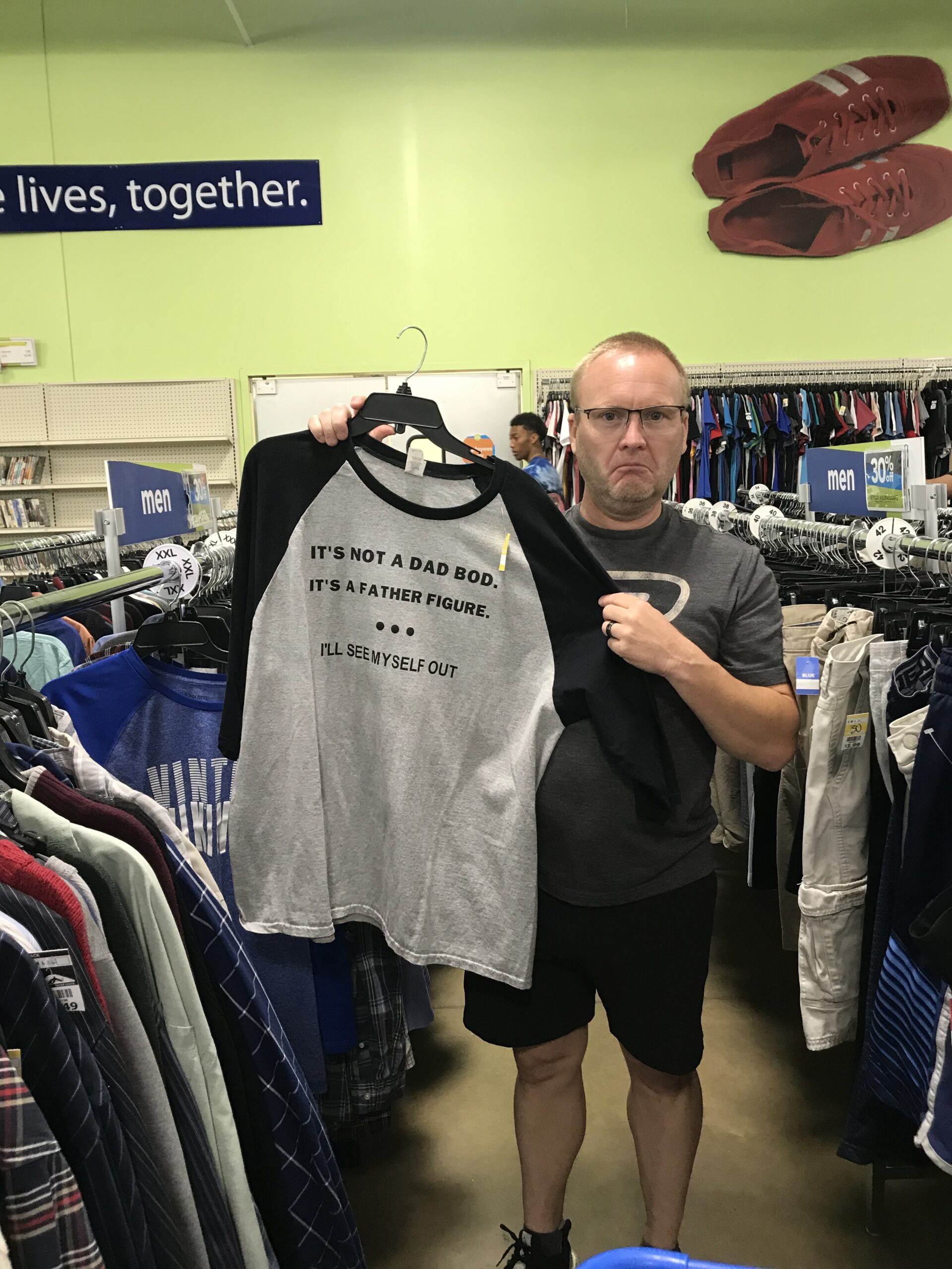 Man standing in a thrift store holding up a gray and black shirt that reads, ‘It’s not a dad bod. It’s a father figure. I’ll see myself out,’ with a humorous, unimpressed expression.