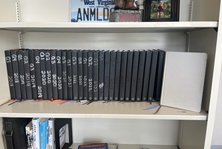 A neatly organized bookshelf displaying a row of black quarterly journals labeled from 2018 through 2023, each marked “Q1” through “Q4.” A light gray journal stands at the far right. On the shelf above sits a decorative West Virginia license plate reading “ANMLDR,” a small statue, and a framed photograph of two people. The lower shelf holds several books, including titles on management and leadership.
