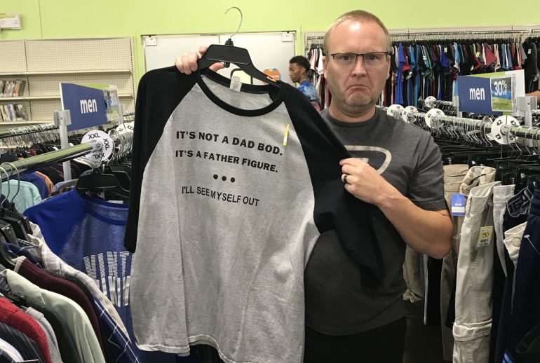 Man standing in a thrift store holding up a gray and black shirt that reads, ‘It’s not a dad bod. It’s a father figure. I’ll see myself out,’ with a humorous, unimpressed expression.